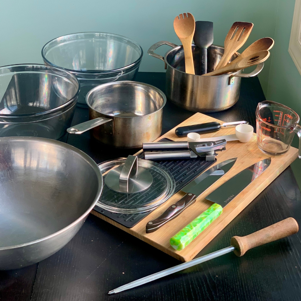 essential basic cooking tools for beginners laid out on a table, image shows bowls, pots and lid wooden utensils and cutting tools plus small items like measuring spoon, can opener and knife sharpening steel rod.