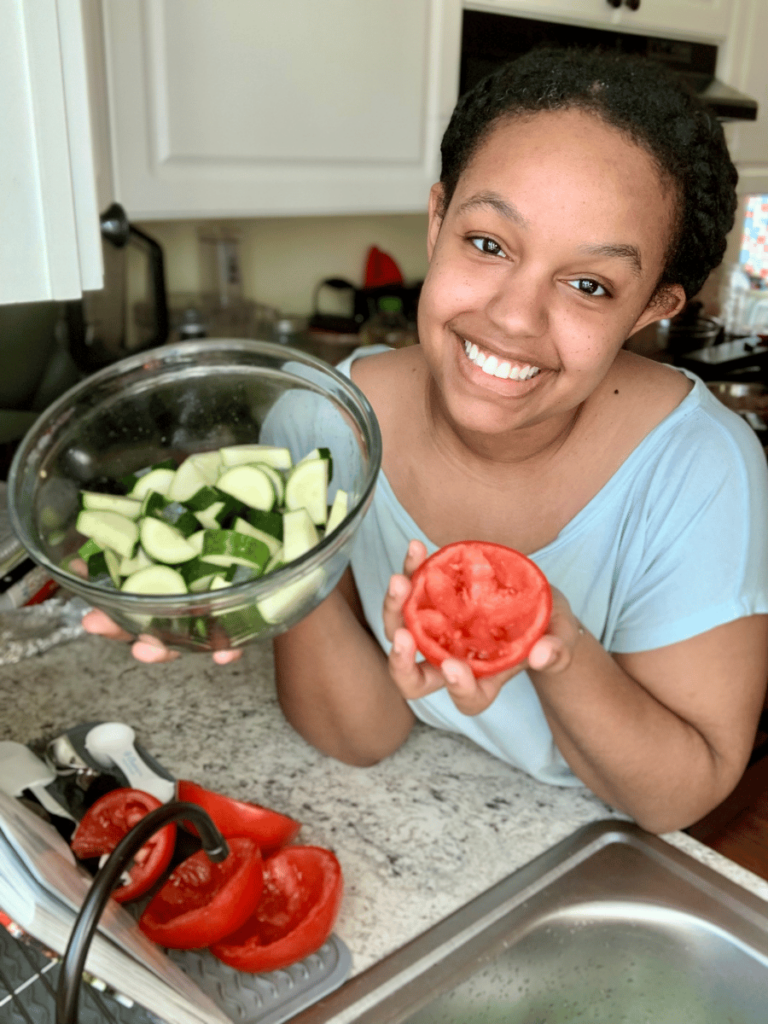 brown girl smiling holding a bowl of sliced zucchini in one hand & a cut & cored tomato in the other hand