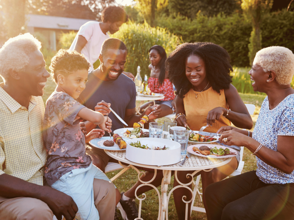 family members sitting around tables in a yard or garden outside in the sunshine smiling & eating good food