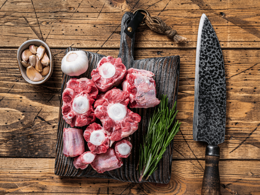 raw oxtails lie on a wooden cutting board with a sharp knife, some herb and garlic in a small dish