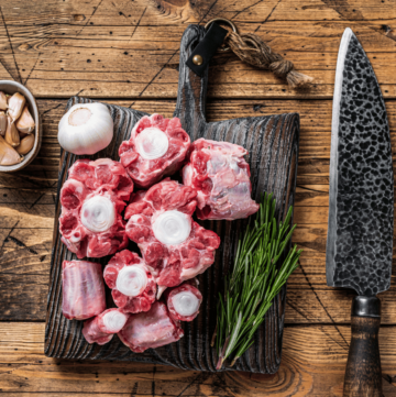 raw oxtails lie on a wooden cutting board with a sharp knife, some herb and garlic in a small dish