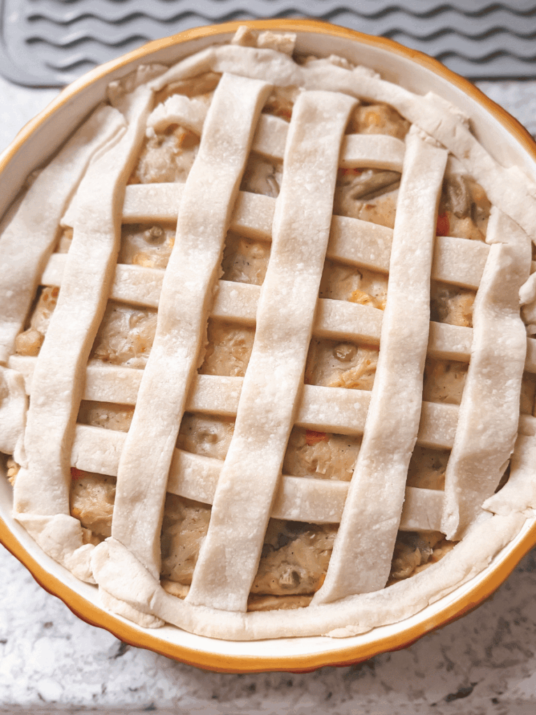 circular ceramic pie pan filled with chicken pot pie filling with a lattice-patterned pie crust topping. Unbaked homemade chicken pot pie is rustic, with imperfect lattice lines and edges.