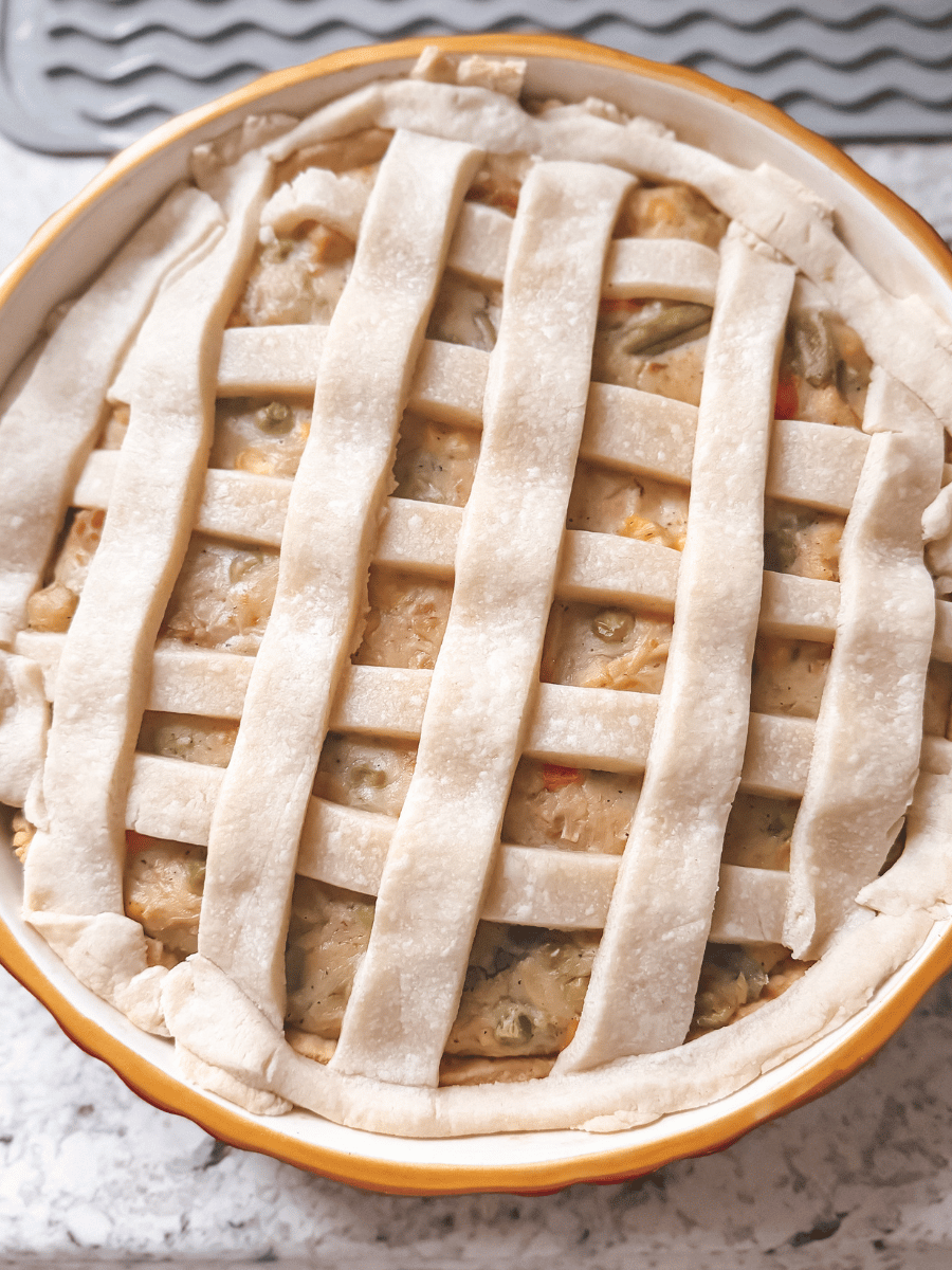 circular ceramic pie pan filled with chicken pot pie filling with a lattice-patterned pie crust topping. Unbaked homemade chicken pot pie is rustic, with imperfect lattice lines and edges.