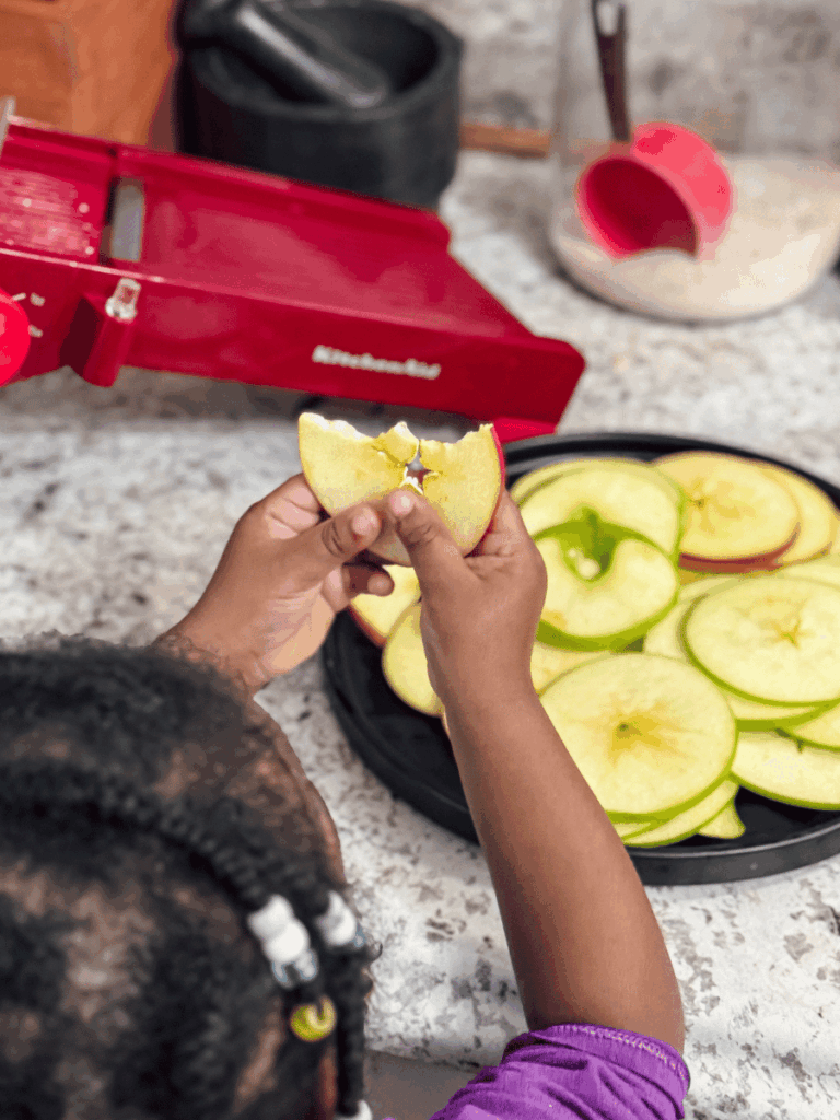 small brown girl toddler facing away from the camera holds up a thinly sliced apple ring that has the bite marks of a child. A plate of more thinly sliced apple rings sits in front of her with a red Kitchen Aid mandolin in the background.