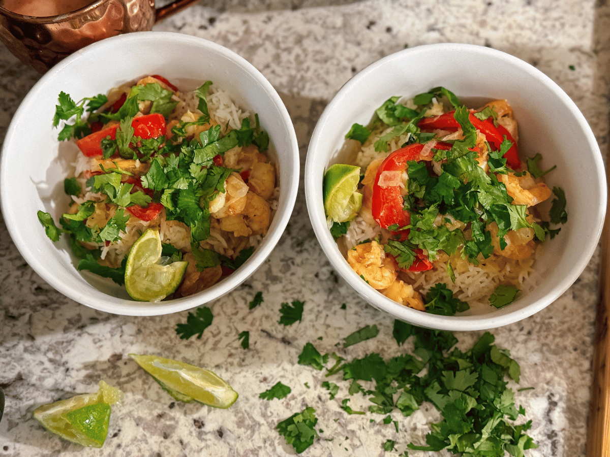 two white bowls sit on a counter and are filled with white rice, pink shrimp, red bell peppers, lime wedges & bright green cilantro. Cilantro is also piled on the counter with more lime wedges.