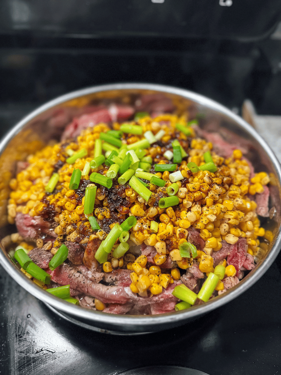 a pan of raw to medium rare beef sits underneath a bright yellow mound of corn kernels & is sprinkled with vibrant green onion slices and dark brown spices and pepper. This recipe for beef pepper rice from Japan is a true comfort family meal.