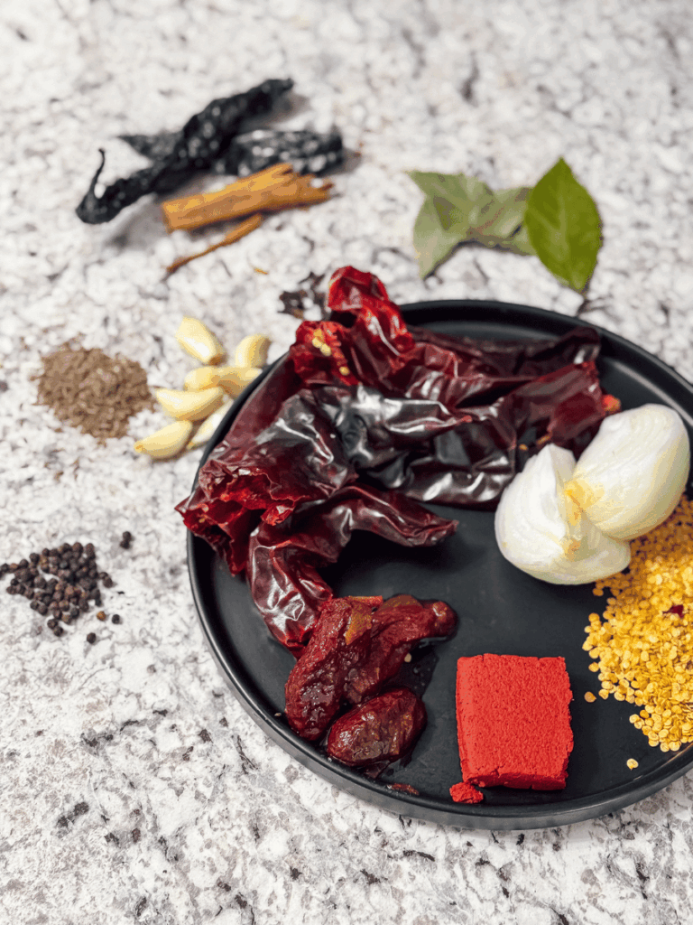 a round plate full of ethnic chiles, spices & pastes. Various cooking seeds, cinnamon sticks, bay leaves, onions and garlic are also spread around the plate and counter.