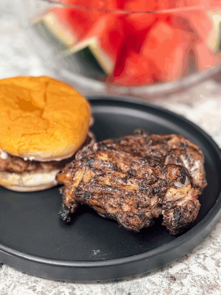 a grilled Caribbean jerk marinaded chicken thigh rests on a black, circular ceramic plate next to a hamburger and bowl of red and green watermelon.