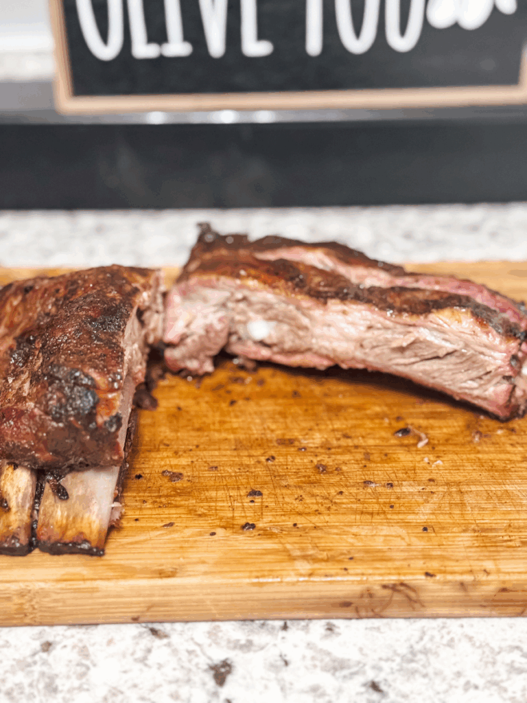 a half rack of smoked or oven-broiled BBQ ribs sit on a wooden cutting board on a countertop. They are visibly juicy & look well-seasoned. The inner meat is richly browned although a light hint of pink remains. Instant pot baby back or pork spare ribs make a delicious comforting family meal on a weeknight.