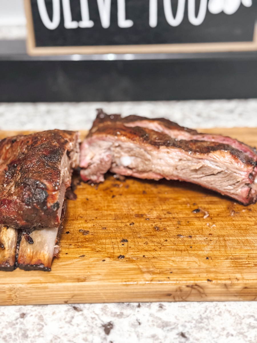several smoked BBQ ribs sit on a wooden cutting board on a countertop. They are visibly juicy & look well-seasoned.
