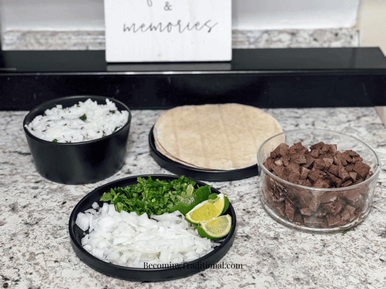 a homemade feast of tacos de lengua is laid out on a countertop. There is a transparent glass bowl filled with browned cow tongue or beef tongue meat, two small black circular ceramic plates - one with flour tortillas & the other with diced white onions, bright green cilantro & lime wedges & a small circular black ceramic bowl with white rice speckled with green cilantro.