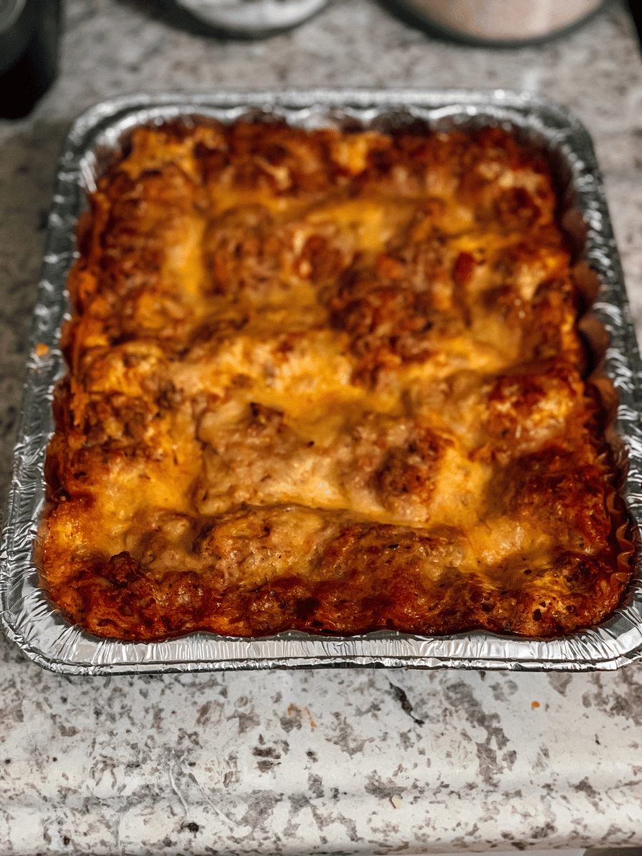 an aluminum pan filled homemade lasagna bolognese with a bechamel sauce sits on a countertop. The cheese layer on the top of the lasagna is bubbly & browned.