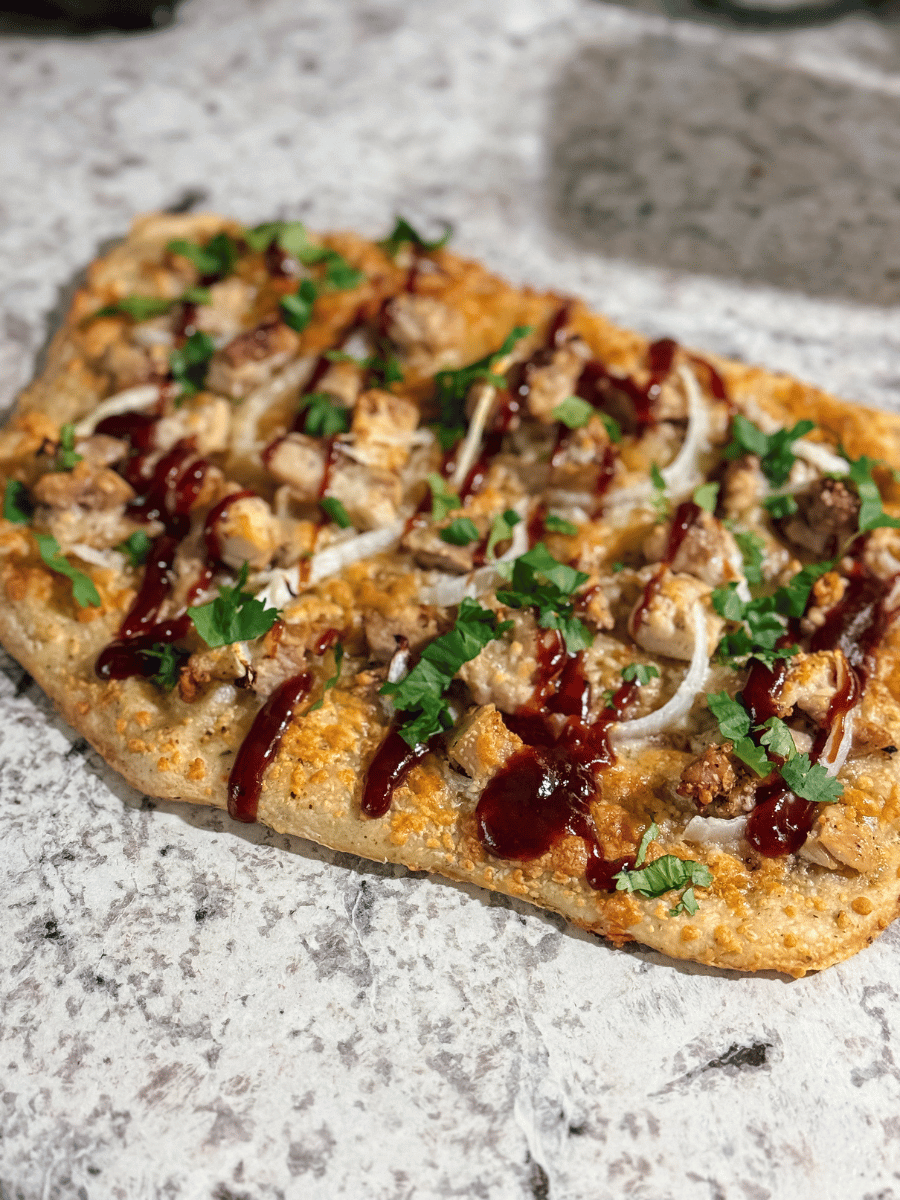 a homemade flatbread pizza lies on a countertop. It is topped with diced seasoned chicken, sliced white onions, green cilantro. Drizzled with a rich brown honey BBQ sauce & sprinkled with cheese that has turned golden brown in the oven. The whole flatbread is cut into even portions to share at the family dinner table.