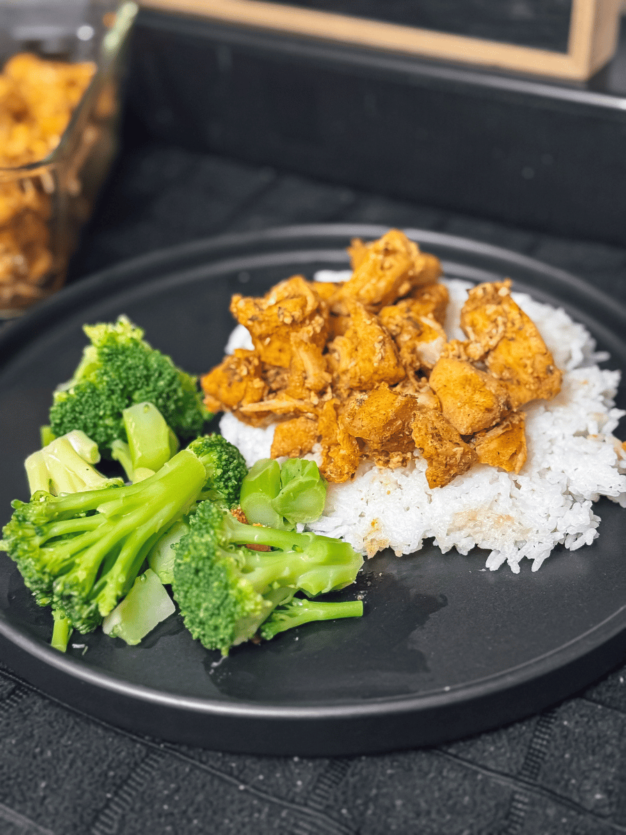 a small circular, black ceramic plate sits on a black towel and has a serving of bright green broccoli. There is also some white rice topped with seasoned browned diced chicken.